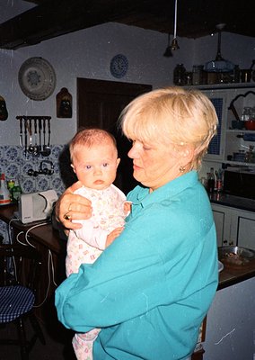 A woman in a teal apron holds a baby in a vintage kitchen with retro decor, including wall clocks, floral-patterned dishware,...