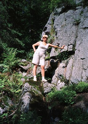 Woman in 1970s-style outdoor wear poses confidently on rocky terrain surrounded by ferns and moss, likely in a mountainous or...