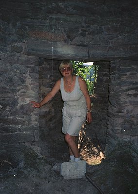 Woman in 1970s-style light-colored jumpsuit poses in a rustic stone archway, sunlight streaming through. Stonework suggests h...