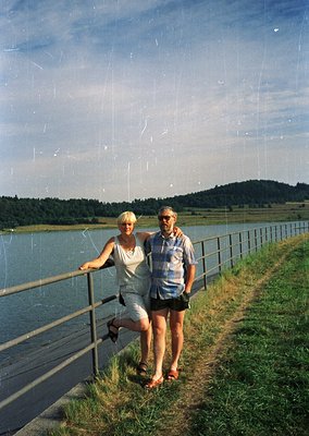 Couple posing on a lakeside promenade with metal railings, 1970s retro fashion—women in light linen dress, men in striped shi...