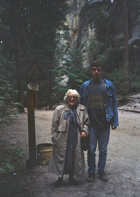 A man and woman pose on a forested trail near a wooden signpost with directional arrows and a metal trash bin. The woman wear...