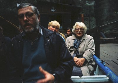 Vintage group shot of three adults on a blue watercraft, likely a ferry or boat tour. Man in foreground wears round glasses, ...