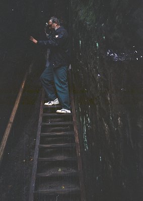A person stands on a narrow, wet metal staircase descending into a dark, flooded tunnel or mine shaft, holding a camera up to...