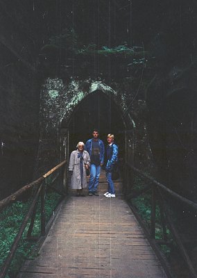 Three individuals stand on a wooden walkway leading into a dimly lit cave entrance, likely a historic or tourist site. The ma...