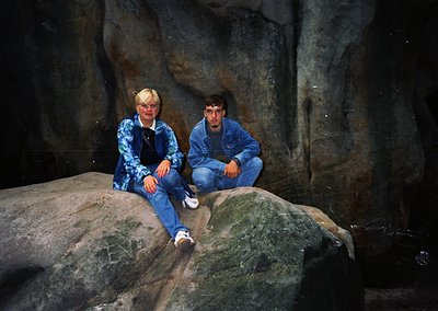 Two individuals pose on moss-covered rocks in a dimly lit cavern, wearing 1990s-style denim jackets and jeans. The rough ston...