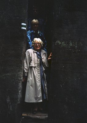 Vintage black-and-white photo of two women in a dimly lit, narrow stone corridor. The woman in the foreground wears a long co...