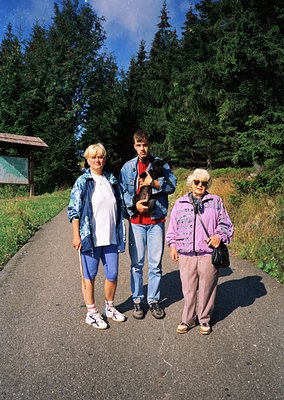 Three individuals pose outdoors on a paved path surrounded by dense forest. The woman on the left wears a denim jacket and cr...
