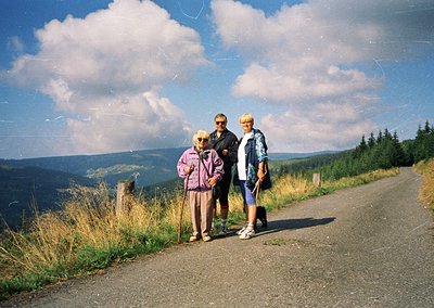 Three adults pose on a paved mountain road, surrounded by forested hills under bright skies. The elderly woman wears a purple...