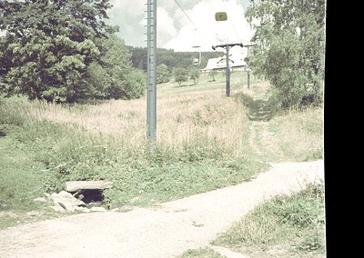 Rural dirt road bordered by overgrown grass and trees, with a utility pole and power lines. A small stone drainage structure ...