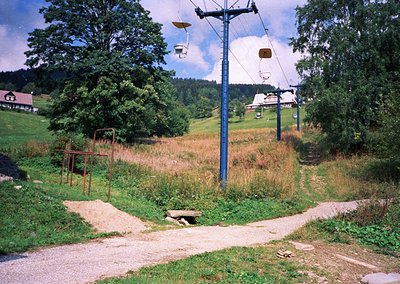 Vintage alpine chairlift station with mid-century design, featuring rusted metal benches and a winding dirt road. Green fores...