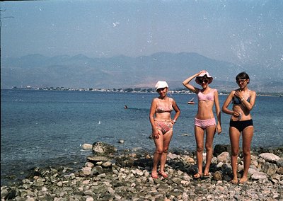 Vintage seaside photo featuring three women in 1960s-style swimwear posing on rocky shore. Bright sunlight reflects off water...