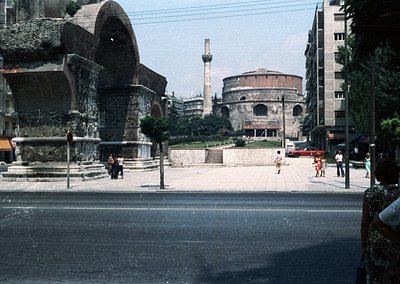 Roman-era amphitheater ruins dominate foreground, blending with mid-20th-century urban infrastructure. Circular stone seating...