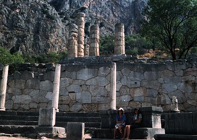 Ancient Greek ruins featuring Doric columns and stone foundations, set against rugged mountainous terrain. Two individuals si...