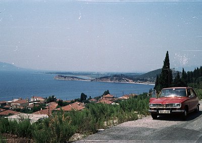Vintage red sedan parked on a coastal hillside overlooking a serene bay with distant islands, framed by lush greenery and Med...