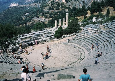 Ancient Greek amphitheater with tiered stone seating and central performance area, surrounded by scattered ruins and greenery...