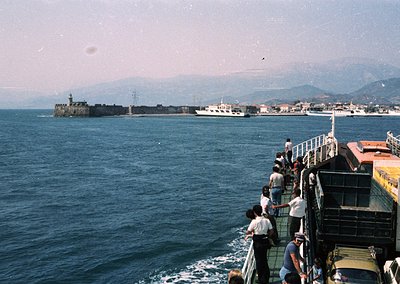 Passengers gather on a ferry’s deck, observing a coastal fortress and port town. The structure features a prominent lighthous...
