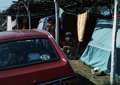 Vintage red car with "CS" sticker parked beside makeshift tents and tarps in a campsite setting. Mid-20th century outdoor liv...