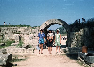 Three women in 1970s summer attire pose under a Roman-era stone archway, likely part of an ancient amphitheater or public com...