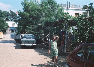 Vintage street scene featuring a woman in a green sleeveless dress posing near lush greenery, flanked by classic Soviet-era c...