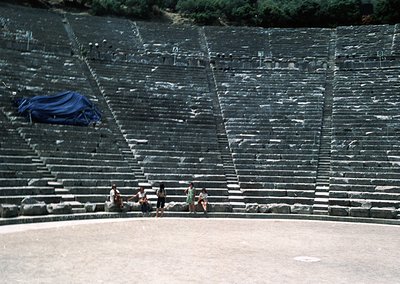 Ancient Greek-style amphitheater with tiered stone seating, partially covered by a blue tarp. Four individuals sit on the low...