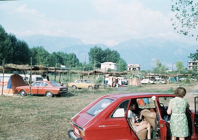 Vintage outdoor gathering in a rural setting, featuring 1970s-era cars and makeshift tents. A red hatchback dominates foregro...