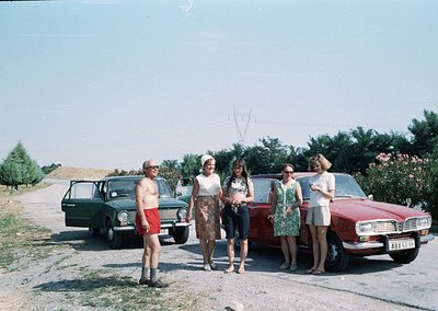 Vintage group photo by rural road, 1970s Eastern Europe. Five individuals pose beside two classic cars: a black Lada and a ma...