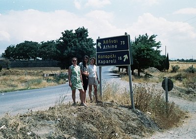 Two women pose beside a Greek road sign (1970s) pointing to Athens (73km) and Kaparelli (16km). Rural countryside with dry ve...