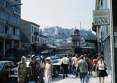Busy urban street in Athens, Greece, with Acropolis visible in background. Mid-20th century clothing and vintage cars suggest...
