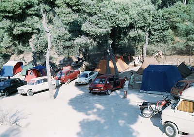 Aerial view of a mid-20th-century camping site with vintage vehicles (1960s-1970s) parked among tents and caravans under dens...