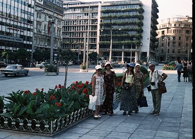 Vintage urban scene featuring five pedestrians in 1960s-era fashion—wide-brim hats, floral dresses, and structured coats—posi...