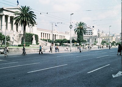 Neoclassical building with grand columns and steps in urban setting, flanked by palm trees. Mid-20th century street scene wit...