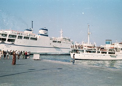 Mid-20th century ferry port scene: Two large passenger ships ("Kantia" and "ANEX") docked at a bustling pier with crowds boar...