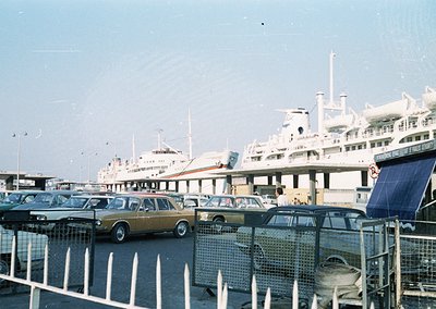 Vintage ferry port scene with mid-20th century passenger ships docked. Yellow classic car (likely a 1970s Ford Escort) parked...