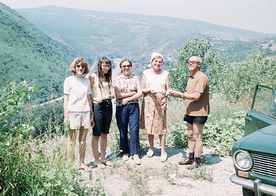 Five individuals pose outdoors in mid-20th-century attire (1960s-1970s) on a grassy hillside, framed by a vintage vehicle. Th...