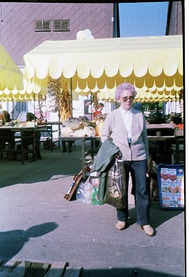 Vintage outdoor market scene featuring an elderly woman in a purple wig, sweater, and apron carrying a bag and broom. Yellow ...