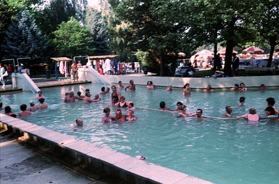 Vintage outdoor swimming pool filled with people in swimsuits, likely mid-20th century. Lush greenery surrounds a shallow, re...