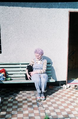 Mid-20th century woman in overalls and striped sweater sits on a wooden bench beside a light-colored building. She holds a sm...