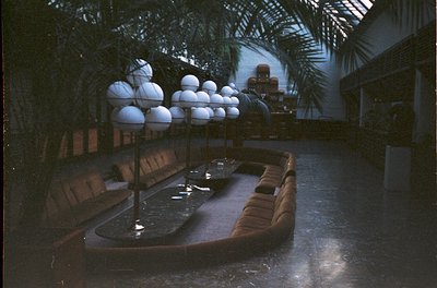 Mid-century modern indoor courtyard with curved leather seating, central shallow pool, and spherical light fixtures. Lush tro...