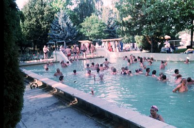 Vintage outdoor swimming pool filled with people, likely from the 1960s–1970s. Concrete edges, shallow diving platform, and l...