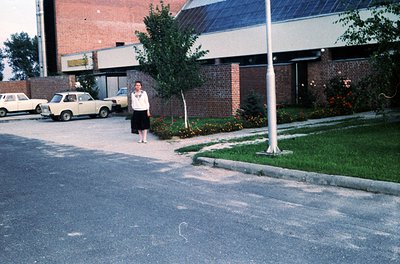 A person in 1970s-era clothing stands on a paved sidewalk beside a brick building with solar panels, likely a public or insti...