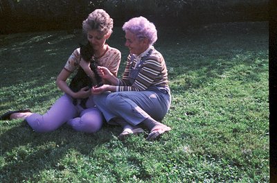 Two women in 1970s outdoor attire—one in purple pants and striped sweater, the other in a patterned blouse—sit on grass holdi...