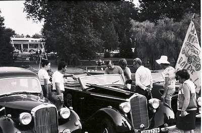 Vintage 1950s-60s gathering featuring classic cars and casual attire. A group of men and women pose near a vintage Mercedes-B...