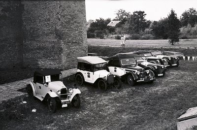 Vintage lineup of classic cars parked beside a stone wall, likely mid-20th century. Open-top models dominate, featuring round...