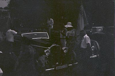 Black-and-white street scene featuring vintage car (license plate "J044229") with passengers in 1940s-50s attire. Crowd gathe...