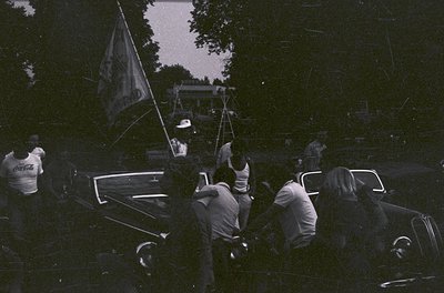 Vintage black-and-white photo of a mid-20th-century outdoor gathering. Group of men in casual 1950s-60s attire (white shirts,...