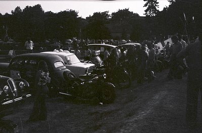 Vintage black-and-white gathering of classic cars and motorcycles in a rural outdoor setting, likely mid-20th century. Packed...
