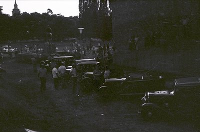 Black-and-white street scene featuring vintage cars (1940s–1950s) and a crowd gathered around a central event. Classic Americ...