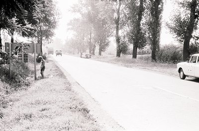 Mid-20th century rural road scene with light dusting of snow. A lone figure in winter attire leans against a fence beside a n...