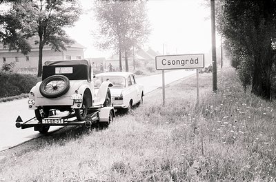 Mid-20th century roadside scene in Csongrád, Hungary. Classic vintage car (1940s-50s) towing a trailer with a newer model veh...