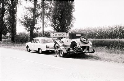 Vintage roadside scene featuring a 1950s-era car towing a classic vehicle with a flat tire. Signpost reads "Csongrád," Hungar...